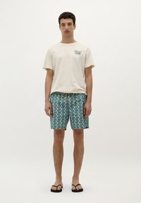 T-shirt en coton beige pour hommes avec impression "Lifeguard Club" ; short de bain à motif teal avec cordon de serrage ; tongs noires.