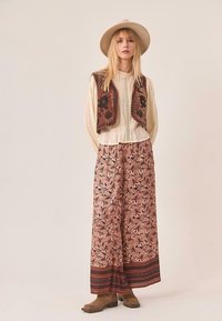 Mujer con blusa beige, chaleco floral marrón, pantalones florales anchos, botas color crema y sombrero de ala ancha beige, de pie frente a un fondo liso.