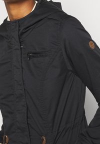 Chaqueta negra con capucha que presenta un bolsillo con cremallera, cintura ajustable con cordón elástico y detalles en marrón. Fabricada con tejido texturizado, diseñada para proporcionar calidez.