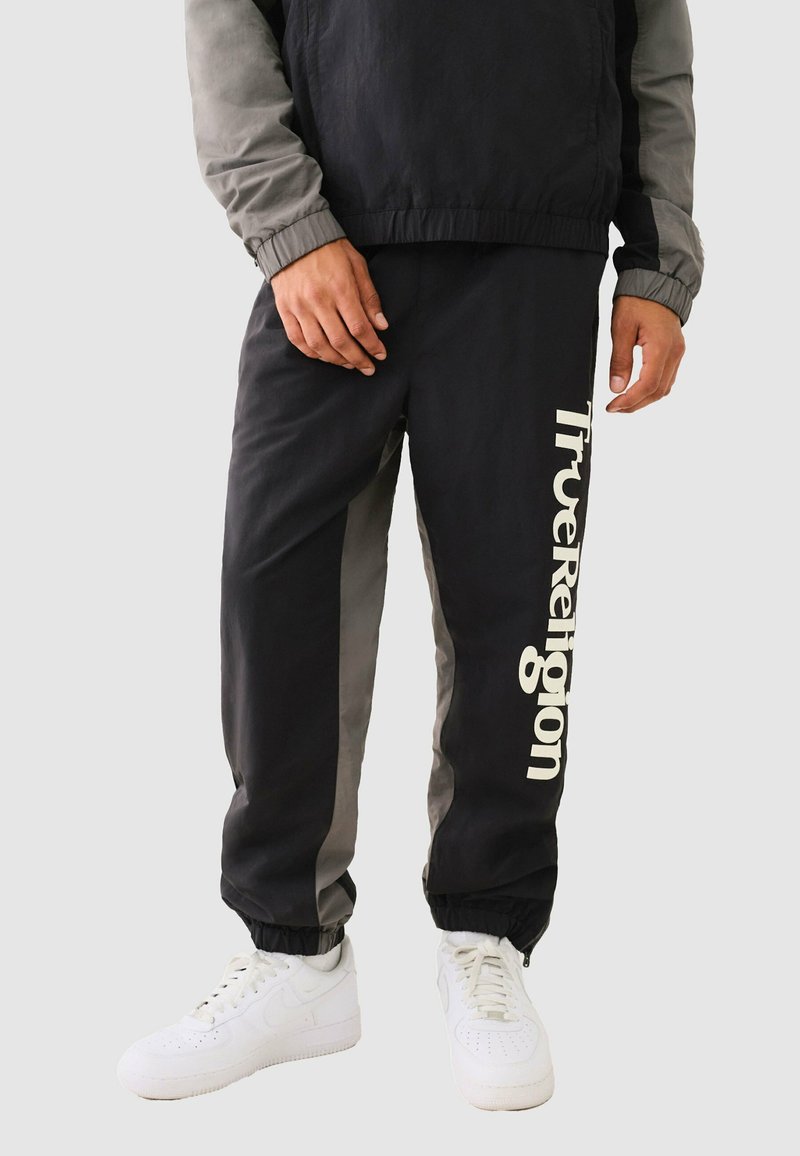Schwarze und graue Jogginghosen mit elastischen Bündchen, die auf der linken Seite ein großes weißes "True Religion"-Logo aufweisen. Glattes, leichtes Material.