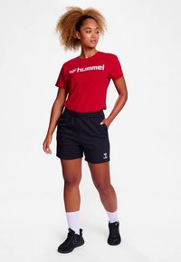 T-shirt rouge à manches courtes avec logo "hummel" blanc, associé à un short de sport noir. Tenue complétée par des baskets noires et des chaussettes blanches montantes.