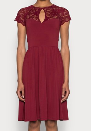 Robe bordeaux avec manches courtes en dentelle, encolure en trou de serrure, corsage ajusté et jupe évasée longueur genou sur une personne debout.