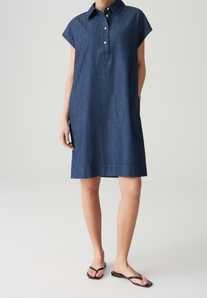 Donna che indossa un vestito di jeans blu scuro a maniche corte, lunghezza al ginocchio, con colletto e bottoni, abbinato a sandali neri con punta aperta.