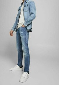 Chaqueta de mezclilla azul claro con dos bolsillos en el pecho, llevada sobre una camiseta blanca y jeans azul claro; combinada con zapatillas blancas.