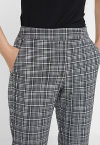 Černobílé kostkované kalhoty s rukama v bočních kapsách, nošené s černým topem zasunutým dovnitř.