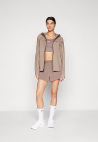 Veste légère marron avec capuche, brassière de sport et shorts marron assortis, chaussettes blanches et baskets blanches. Texture lisse, coupe athlétique.