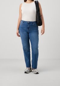 Mujer vistiendo una blusa sin mangas blanca, jeans azules, zapatillas blancas y llevando un bolso negro de hombro frente a un fondo liso.