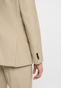 Beige suit jacket and matching trousers with black buttons on sleeve, hand relaxed by side against plain background.