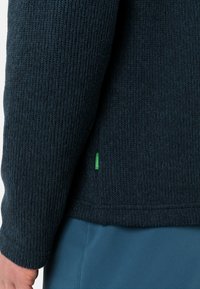 Pulover tricotat de culoare bleumarin, cu o textură canelată, mâneci lungi și un mic etichetă de accent verde la tivul lateral.