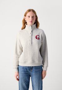 Grå sweatshirt med hög krage, knappslå och ett vinrött broderat bokstav "G" på bröstet. Har ribbade muddar och nederkant.