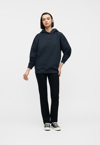 Νεαρή γυναίκα με μαύρη κουκούλα, μαύρο παντελόνι και μαύρα αθλητικά παπούτσια, στέκεται με χαλαρή στάση μπροστά σε λευκό φόντο.