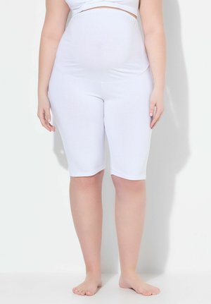 Osoba w ciąży ubrana w białe szorty ciążowe i krótki biały top, stojąca boso na tle jednolitego białego tła.