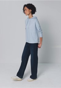 Lyseblå hættetrøje med lange ærmer, kombineret med mørkeblå vidde bukser og hvide sneakers, mod en neutral baggrund.