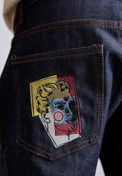 Dark blue denim jeans with contrasting orange stitching. Back pocket features a colorful embroidered patch of stylized faces in varied shapes.