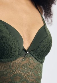 Grön spetsbodysuit med en vadderad, djup urringad bh med blommönster, justerbara remmar och en dekorativ rosett med en kristallaccent i mitten.