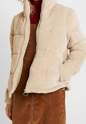 Vrouw draagt een beige corduroy pufferjack gedeeltelijk dichtgeritst over een bruine corduroy rok met zichtbare rits aan de voorkant en een bordeaux coltrui.