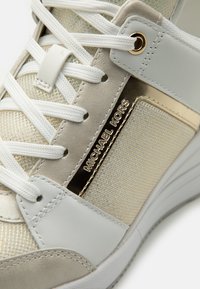 Białe i beżowe sneakersy z siateczkowych i skórzanych materiałów, złotymi akcentami oraz logo "Michael Kors" z boku. Sznurowadła są białe.