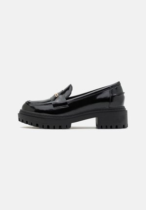 Sort skinnende læderloafer med chunky sål med groft mønster og lav blokhæl, med en lille metallisk spænde på remmen.