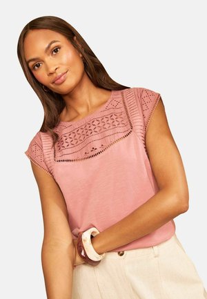 Kvinde med langt brunt hår iført en pink blonde detaljeret ærmeløs top og lyse beige bukser, poserer med to armbånd på håndleddet.