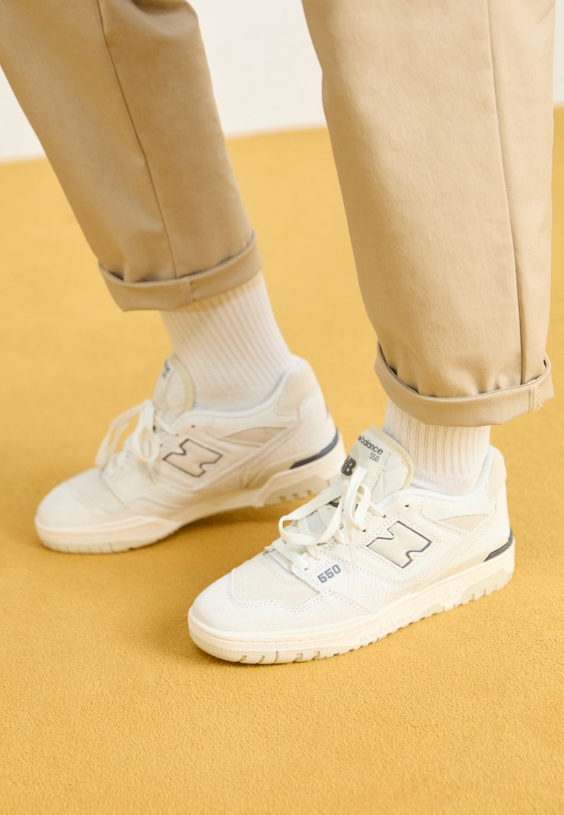 Personne portant des baskets blanches New Balance 550 avec des chaussettes blanches et un pantalon beige retroussé, debout sur un sol jaune.