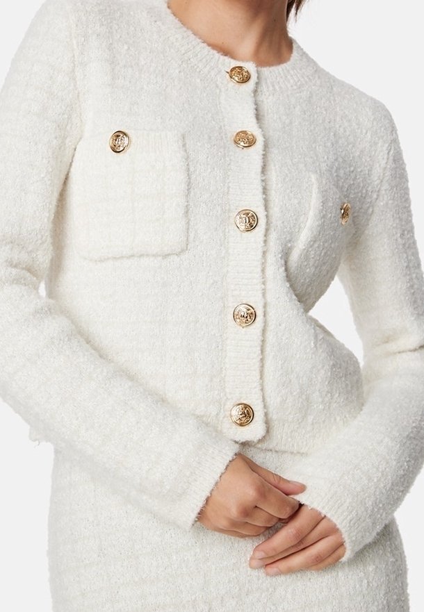 Vit texturerad kofta med guldknappar och två framfickor, med rund halsringning och ribbade ärmslut för extra detalj.