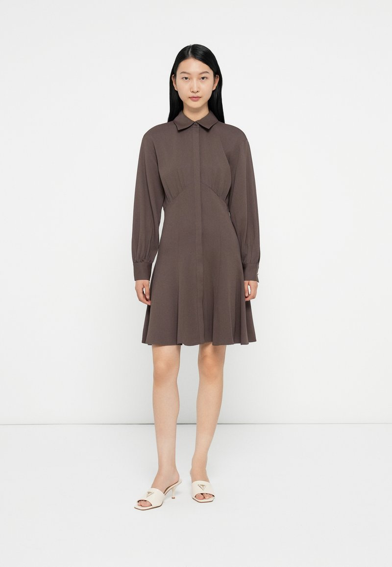 Vestido castanho de mangas longas com gola, corpete justo, saia rodada e fecho de botões. Modelo usa mules de tacão creme.