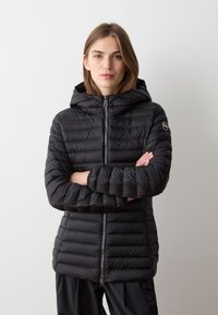 Giacca a vento nera con cappuccio, caratterizzata da una chiusura con zip, design trapuntato e un'etichetta con logo sulla manica sinistra. Realizzata in materiale leggero e liscio.