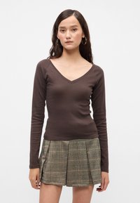 Καφέ μακρυμάνικο μπλουζάκι με V λαιμόκοψη, φτιαγμένο από μαλακό υλικό, σε συνδυασμό με πτυχωτή φούστα καρό σε ουδέτερους τόνους. Διακριτή υφή και εφαρμοστή σιλουέτα.