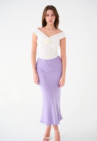 Top off-shoulder bianco con dettaglio annodato al centro, abbinato a una gonna lunga e fluida lavanda. Texture liscia in tutto il capo.