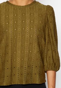 Blusa in verde oliva con texture e maniche a sbuffo, caratterizzata da un design a maglia con motivo e piccoli occhielli aperti. Collo rotondo.