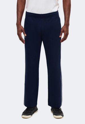 Homme portant un pantalon de survêtement bleu marine avec des bandes blanches sur les côtés et des baskets noires sur un fond clair.