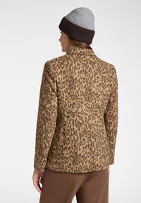 Blazer in stampa leopardata in tessuto marrone e beige, caratterizzato da un design su misura, colletto a punta e silhouette strutturata. Indossato con un berretto grigio.