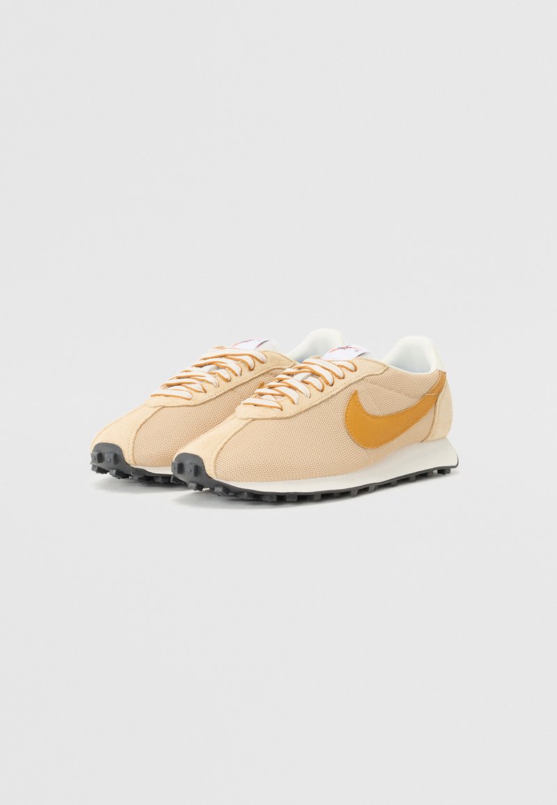 Baskets beiges avec tige en mesh, logo swoosh orange, accents blancs et semelle en caoutchouc noire. Design à lacets, forme classique de coureur.