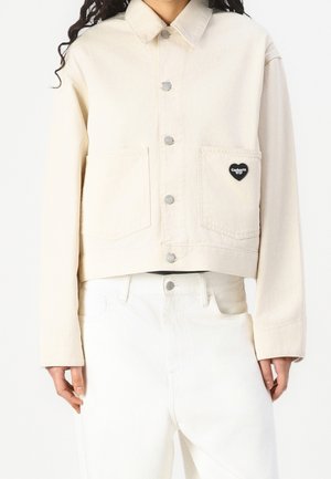 Person wearing a cream-colored cropped jacket with silver buttons and a black heart-shaped patch on the left pocket, paired with white pants.