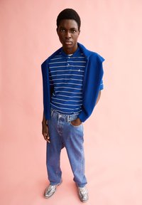 Blau gestreiftes Polohemd mit einem weißen Kragen, kombiniert mit lockeren blauen Jeans. Ein blauer Strickpullover über die Schultern drapiert. Graue Sneakers.