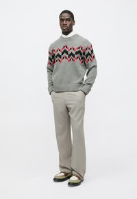 Szürke kötött pulóver geometrikus vörös és fekete mintával, világosszürke nadrággal és színes részletekkel díszített masszív lábbelivel párosítva.