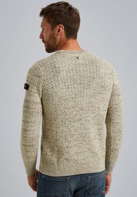 Pull en tricot beige avec un motif texturé, encolure ronde et base côtelée. Présente un petit logo noir sur la manche gauche.