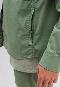 Helle grüne Jacke mit glatter Textur, ausgestattet mit einer Reißverschlusstasche und einer taillierten Passform, schichtweise über einem strukturierten, hellgrauen Hemd.