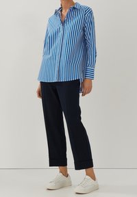 Modro-bílý pruhovaný dlouhý rukáv, volnější střih, s špičatým límcem, v kombinaci s modrými kalhotami a bílými teniskami, stojící na neutrálním pozadí.