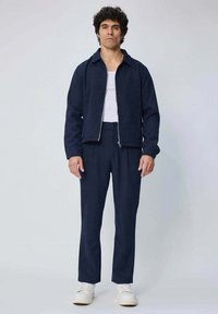 Veste zippée bleu marine avec un design à col, assortie à un pantalon coordonné. Le modèle porte un débardeur blanc et des baskets blanches. Texture lisse.