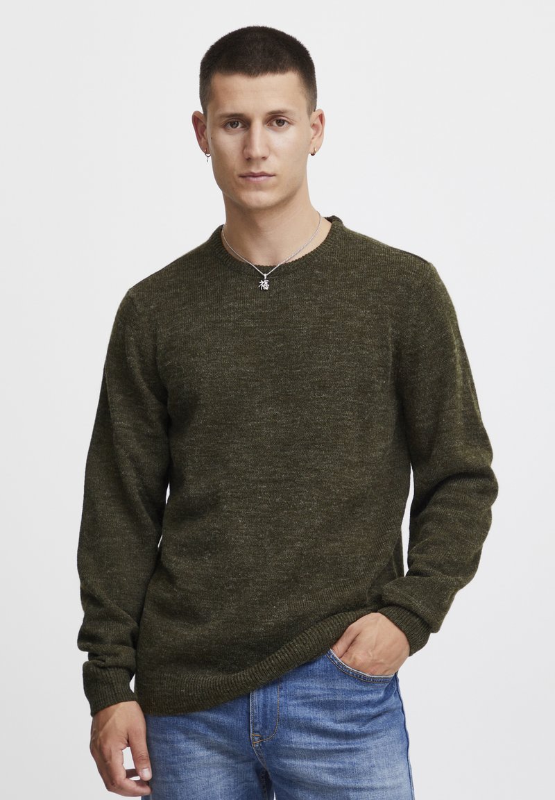 Olivgrüner, gestrickter Pullover mit langen Ärmeln und Rundhalsausschnitt, kombiniert mit blauen Jeans. Die Textur wirkt weich und leicht strukturiert.