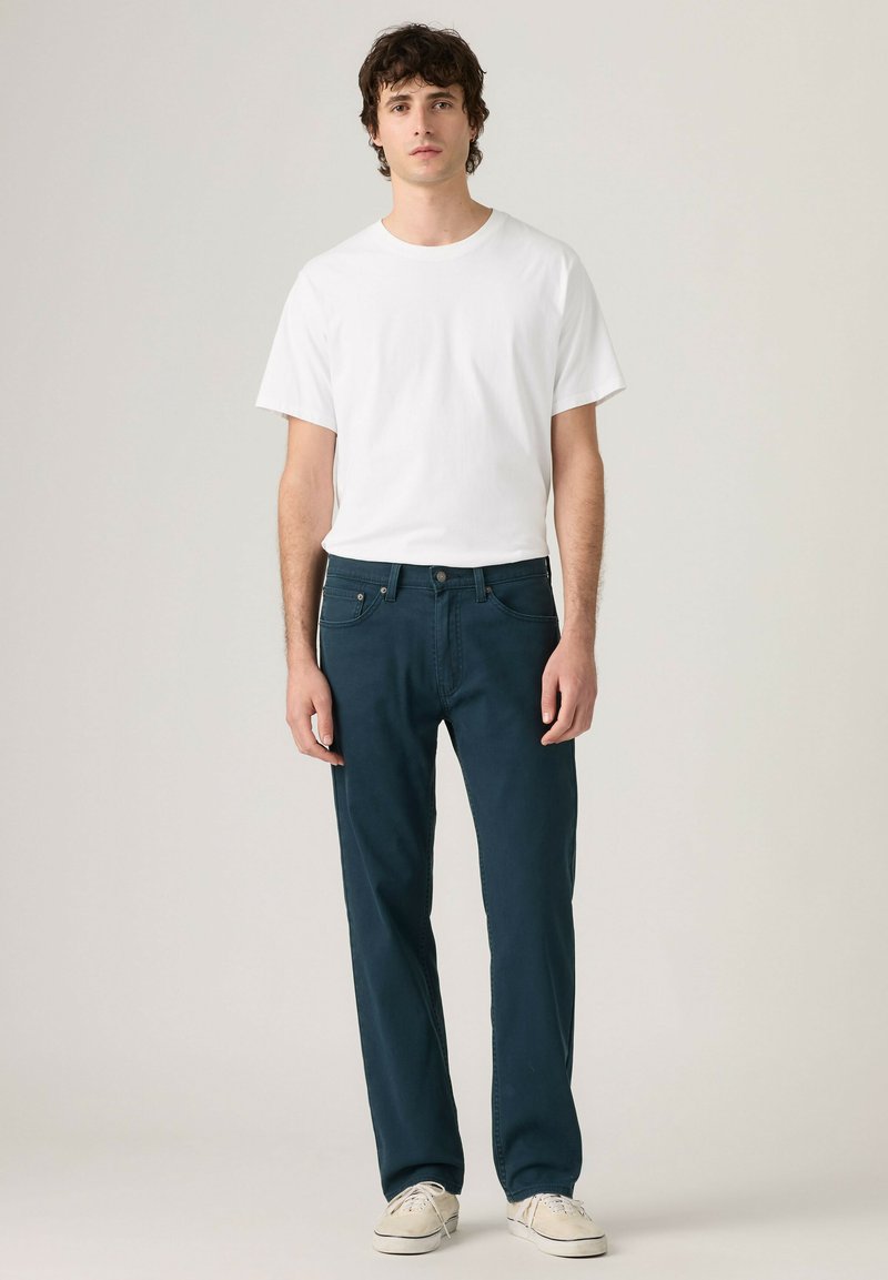 Junger Mann steht und trägt ein schlichtes weißes T-Shirt, dunkelblaue Jeans und hellbeige Sneaker vor einem neutralen Hintergrund.