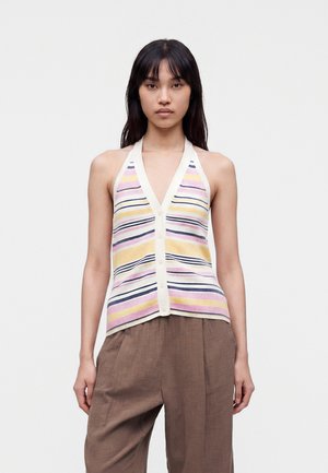 Femme portant un haut sans manches rayé à col halter avec des boutons, associé à un pantalon marron ample, debout devant un fond blanc uni.