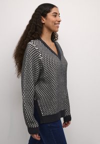 Adīta džemperis ar V veida kakla izgriezumu, ar melnu un krēmkrāsas rūtainu rakstu, ribotiem piedurknēm un apakšmalu, kā arī sānu iedaļām, lai nodrošinātu ērtāku kustību.