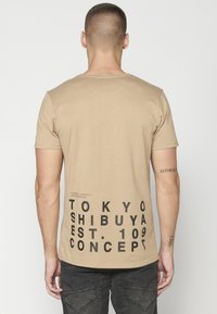 Barna, rövid ujjú póló, hátul nyomtatott fekete szöveggel, mely tartalmazza a "TOKYO SHIBUYA EST. 109 CONCEPT" feliratot és földrajzi koordinátákat.
