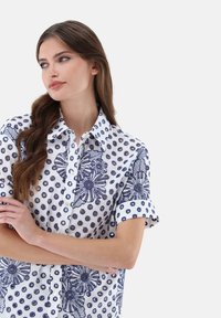 Camicia bianca con ricami blu a fiori e a pois, maniche corte e colletto aperto, realizzata in tessuto leggero.