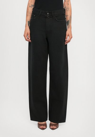 Personne portant un jean noir à taille haute et à jambes larges avec des passants de ceinture et des chaussures à talons ouverts de couleur marron, se tenant devant un fond uni.