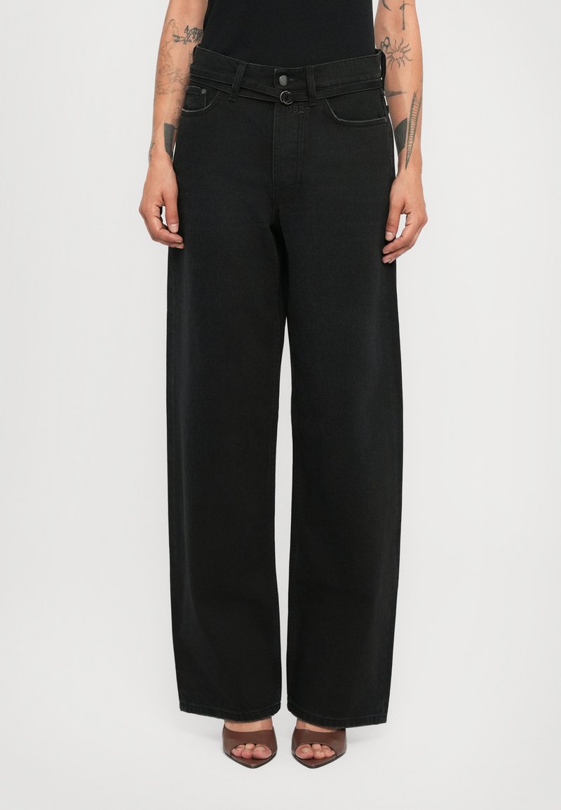 Person wearing high-waist black wide-leg jeans with belt loops and brown open-toe heeled shoes, standing against a plain background.