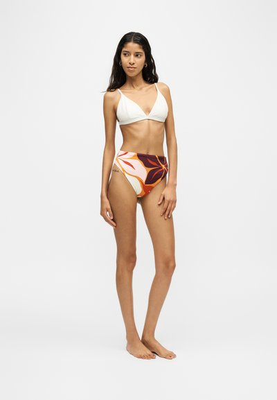 Jeune femme debout, pieds nus, portant un haut de bikini blanc et une culotte taille haute à motif floral orange et violet.