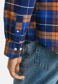 Karierter Hemd mit Blau-, Orange- und Brauntönen, mit Knopfdetails an den Manschetten. Getragen mit Jeans, was einen lässigen Stil zeigt.